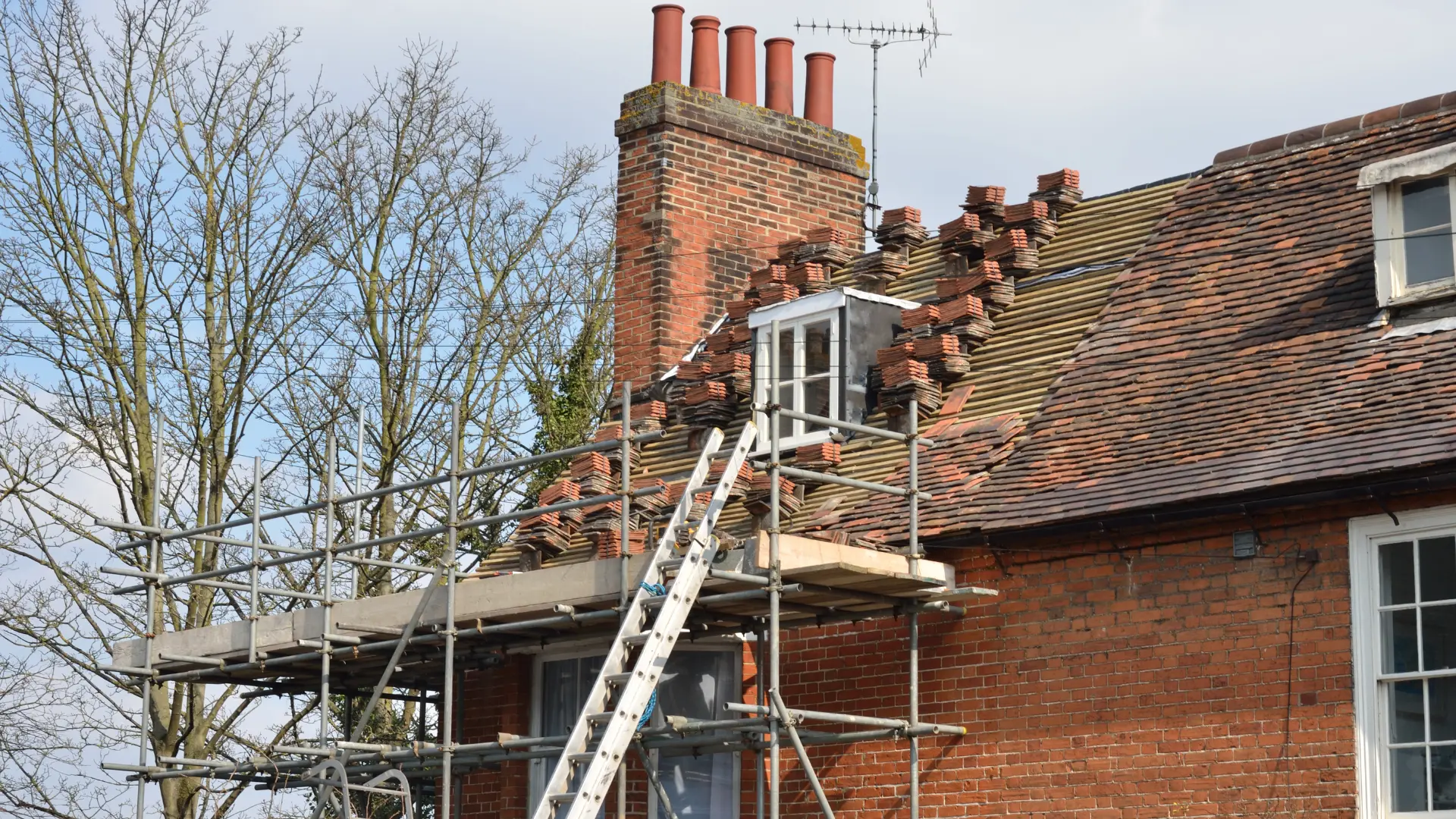 Clay tiles stacked along a stripped roof slope beside a brick chimney during a roof repair